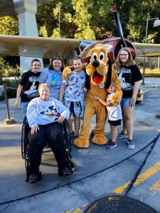 A woman in a wheelchair surrounded by 5 people and Pluto at Disneyland